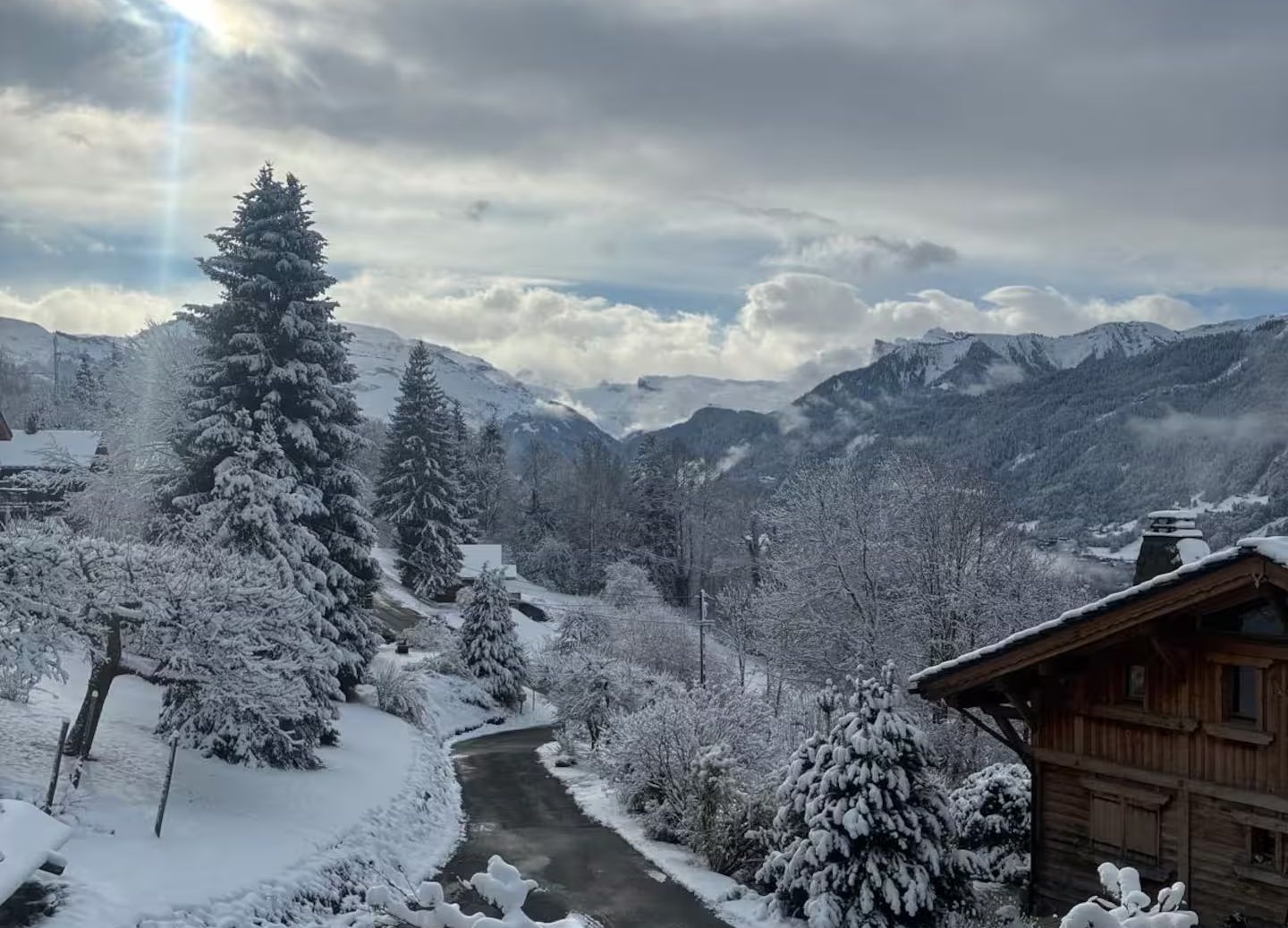 A snowy lane through Verchaix in winter, with a wooden chalet, frosted spruces and the mountains of the Giffre valley beyond